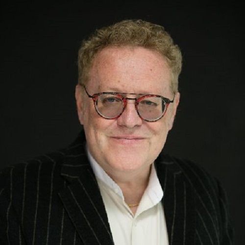 Person wearing glasses and a striped suit jacket against a dark background.